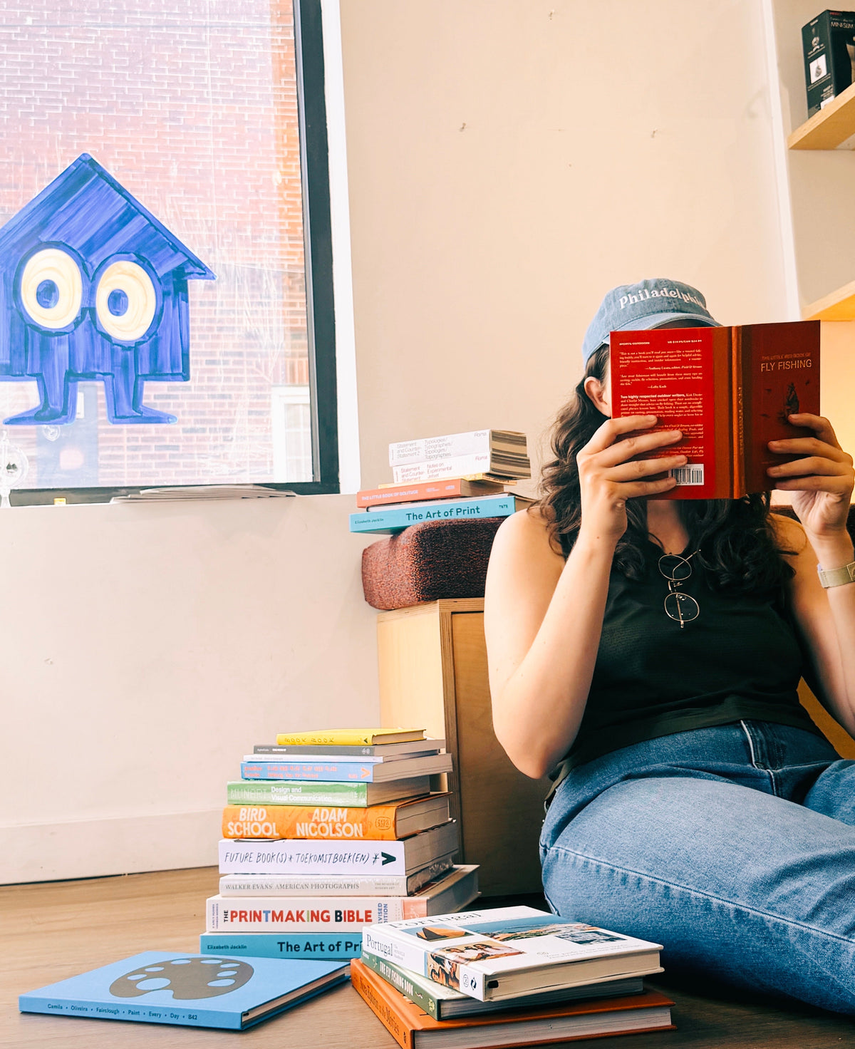 A woman sitting on the floor reads a red book, surrounded by stacks of Another Corner's books in a cozy setting.