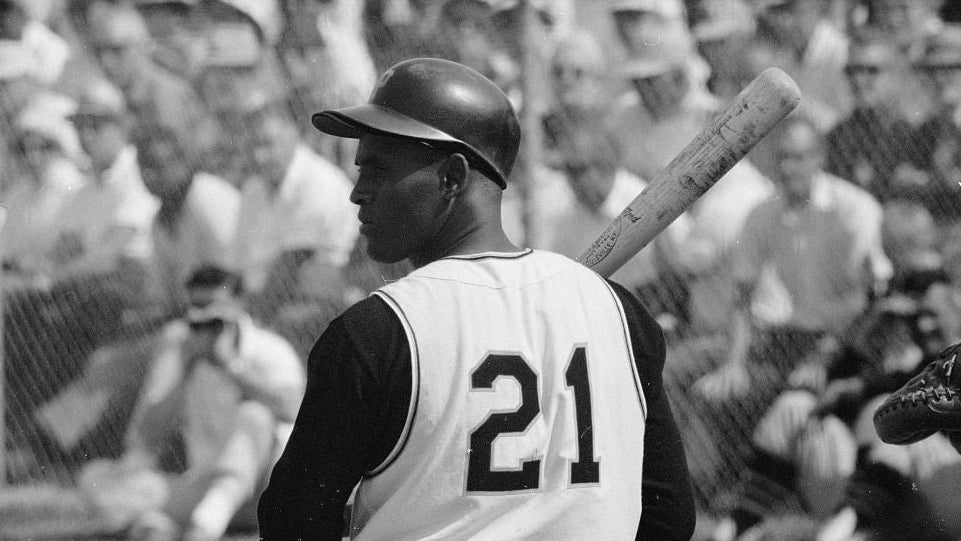 Black and white photograph of Roberto Clemente wearing a helmet and jersey with the number 21, holding a bat over his shoulder on a field with a blurred crowd in the background.